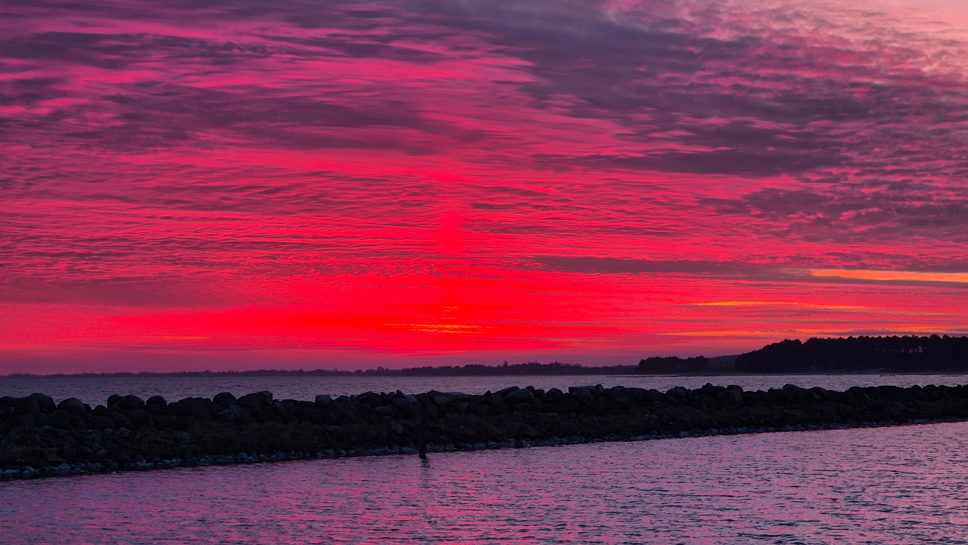 A vibrant pink-red sunset over the calm ocean, with waves gently lapping against the shore and the sky glowing in warm hues.