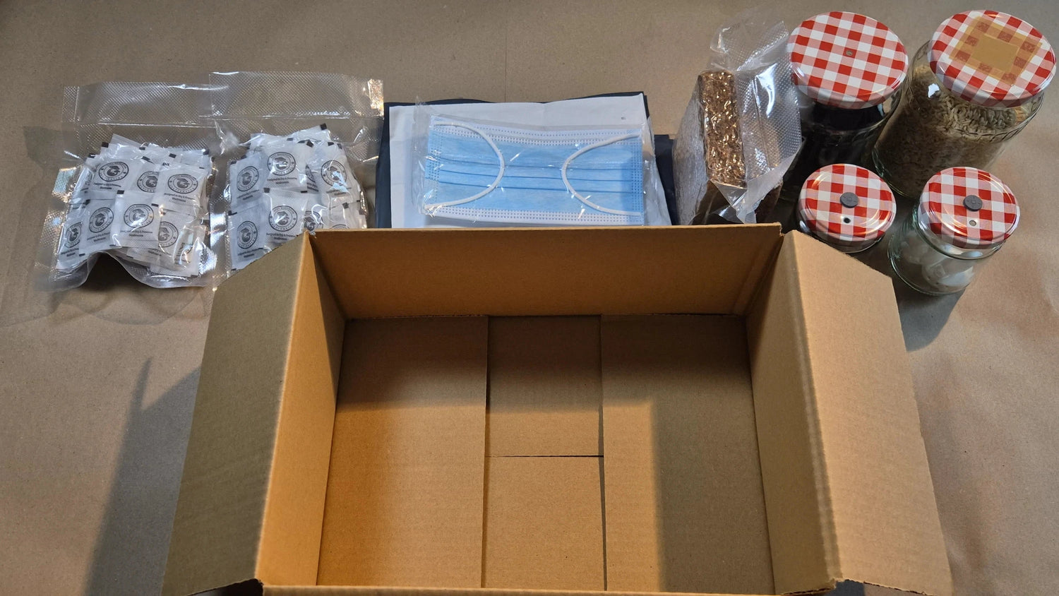 An overhead shot of various cultivation supplies including jars with checkered lids, vacuum-sealed substrate bags, a face mask, and small supplement packets laid out next to an open shipping box.