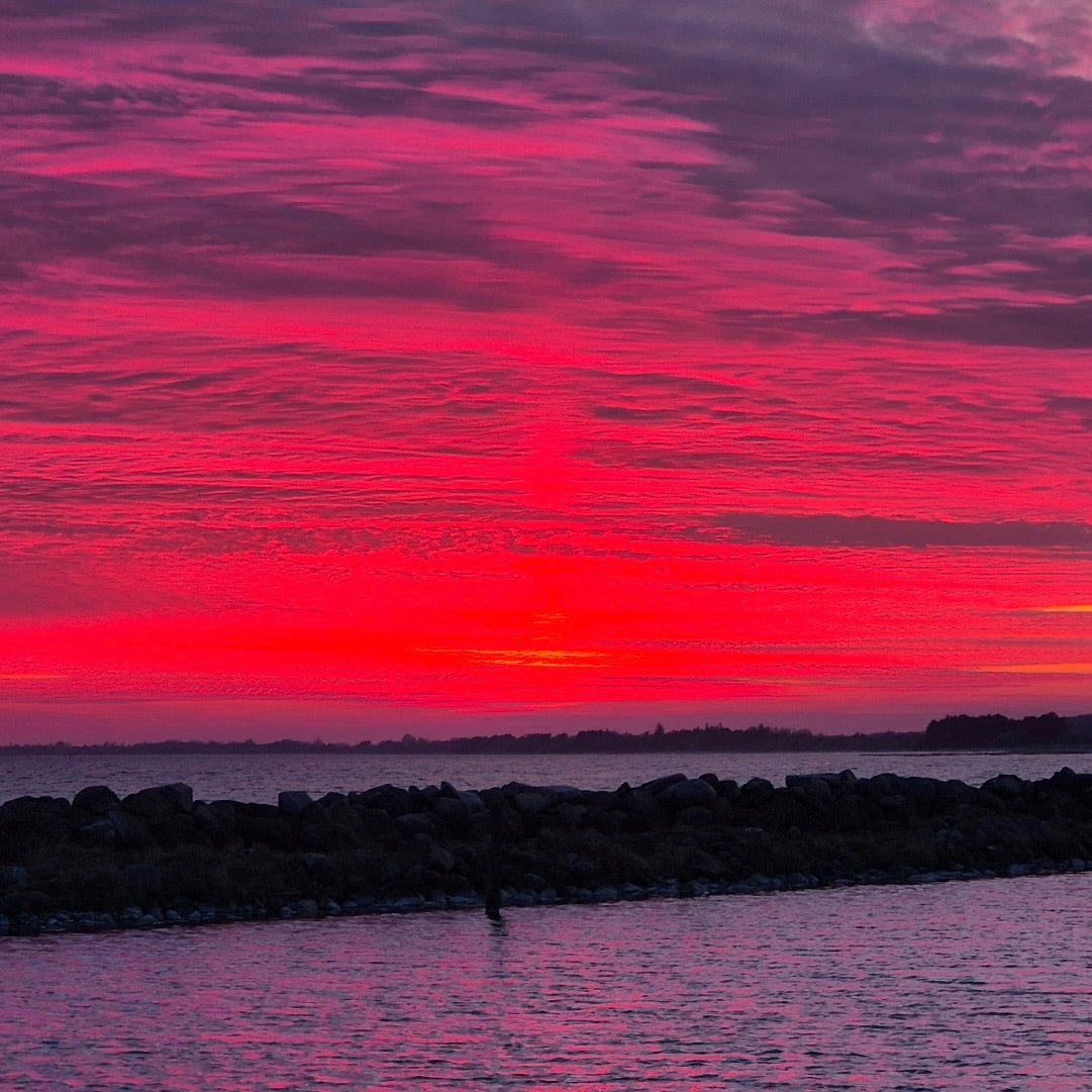 Vibrant red and magenta sunset over a calm ocean with a dark stone breakwater in the foreground.